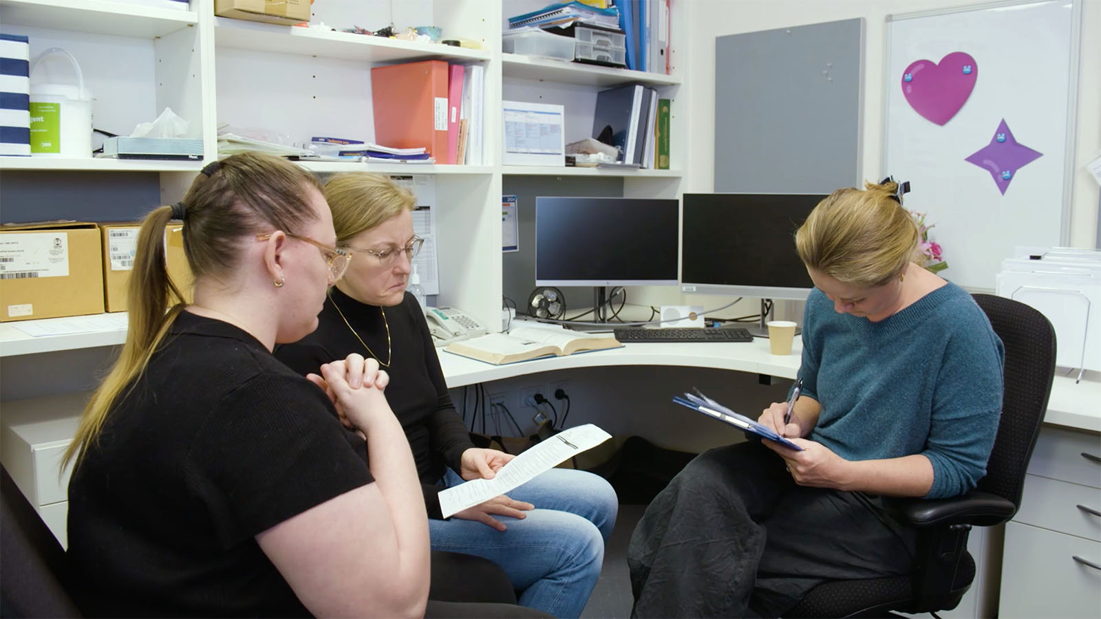 Two women sit in a doctor's office looking at a paper while the doctor writes