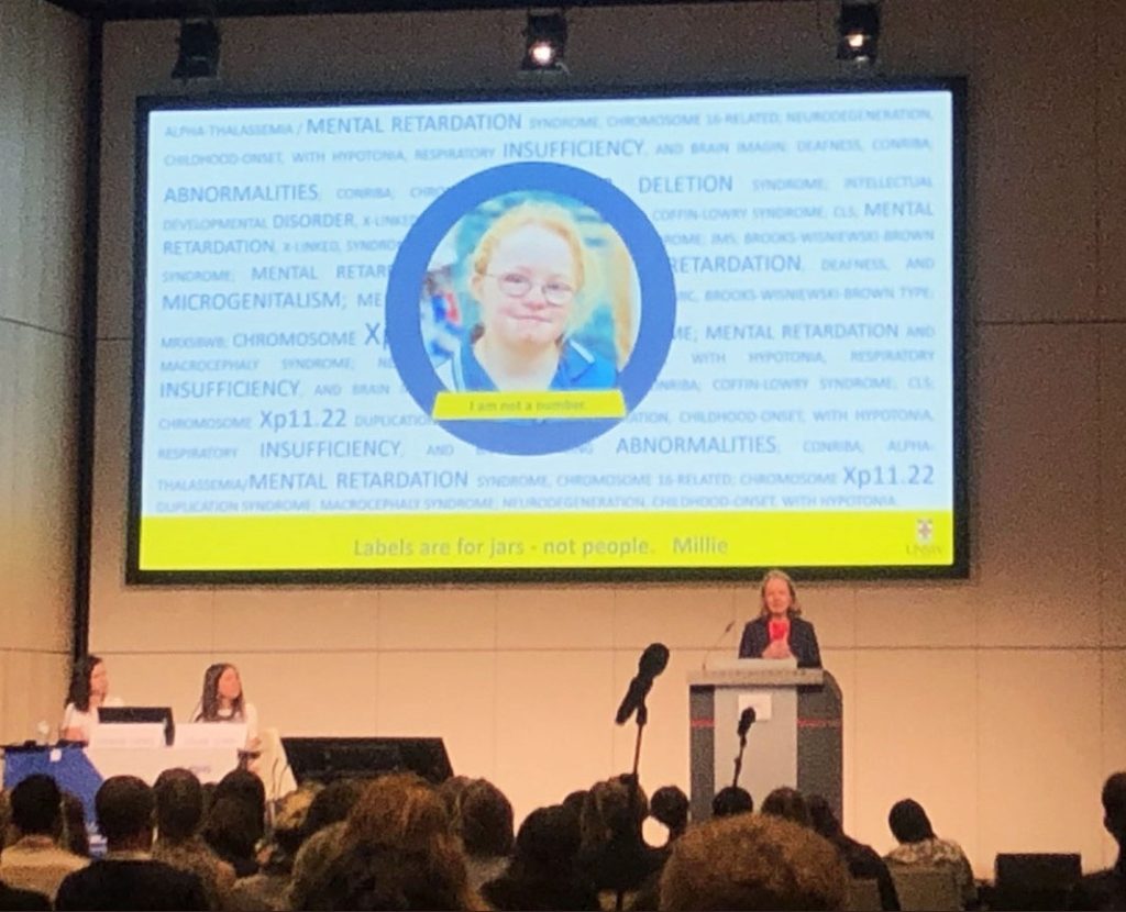 Emma Palmer speaking at a lectern in front of a large screen.