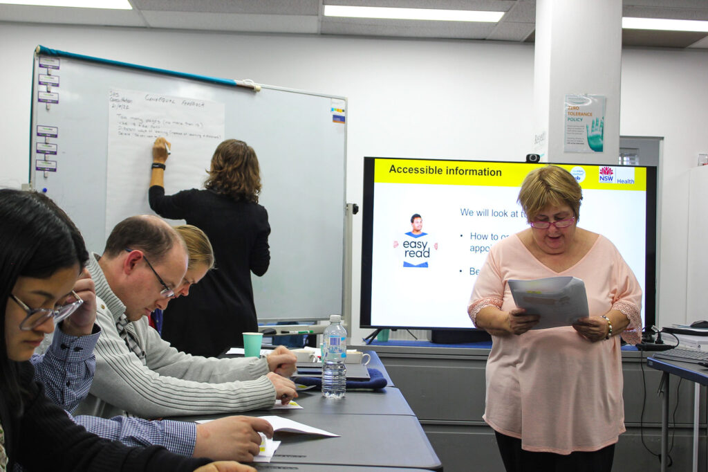 In a conference room, a woman writes on a whiteboard and another reads through a paper while three seated people look through documents